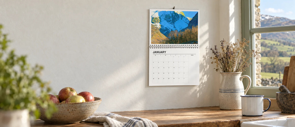Lake District 2027 wall calendar hanging open on a farmhouse kitchen wall beside a window with a view of the fells
