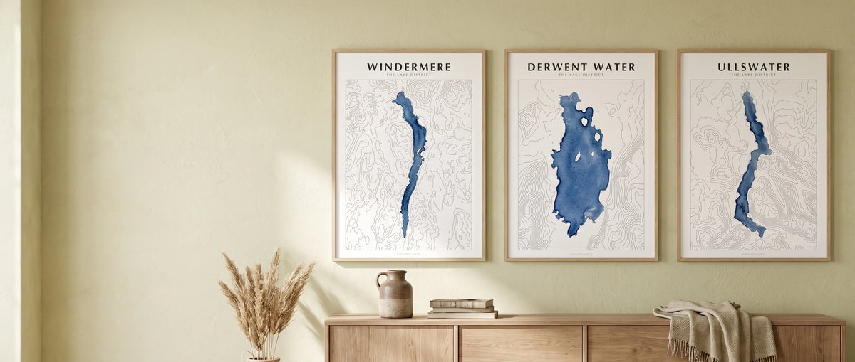 Three framed watercolour lake map posters — Windermere, Derwent Water and Ullswater — hanging in a row above an oak sideboard in a Lake District cottage sitting room